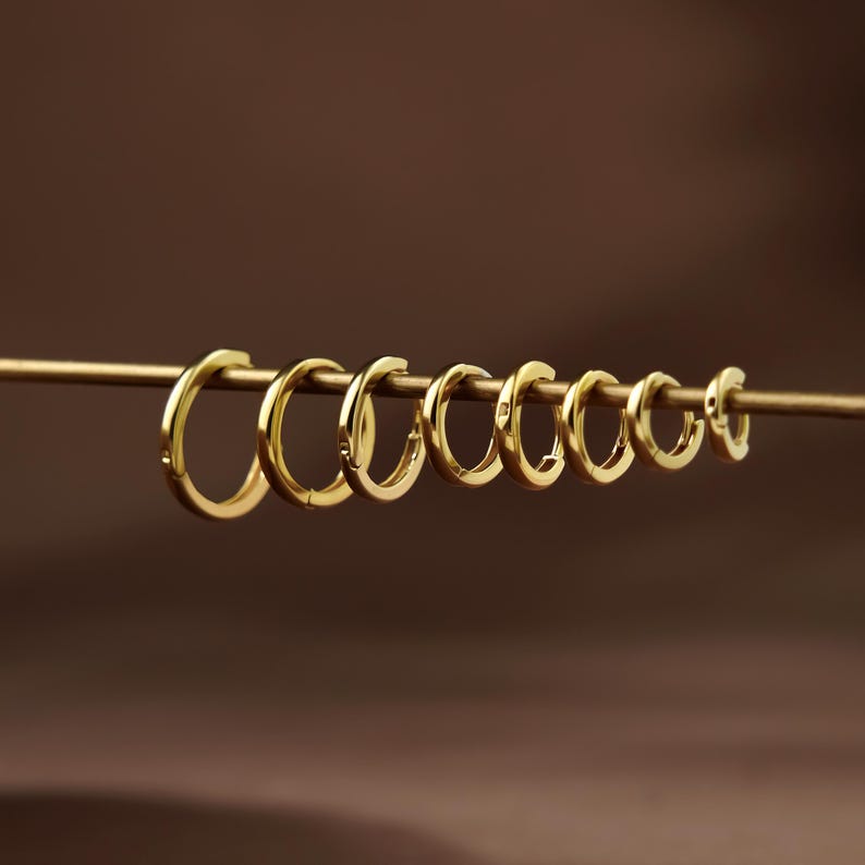 May include: A row of eight polished gold hoop earrings of varying sizes, arranged in a line from largest to smallest on a thin horizontal gold rod. Each hoop features a smooth, circular design with a hinged closure mechanism visible on the side.