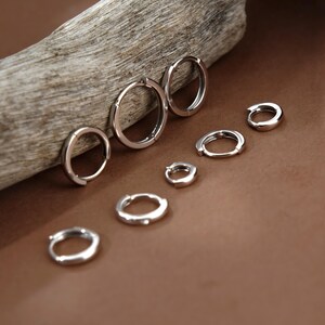 May include: A collection of seven silver-toned hoop earrings of varying sizes arranged in a descending order. The hoops feature a smooth, polished finish and a hinged closure mechanism, displayed in a cluster with some resting on a surface.