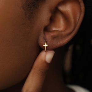 May include: A small gold hoop earring featuring a four-pointed star charm. The star has a polished, reflective surface and is attached to a thin, circular hoop that secures through the earlobe. The design is simple, metallic, and gold-toned.