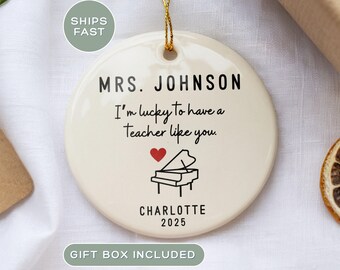 May include: Cream-colored ceramic ornament with gold string, featuring the text "MRS. JOHNSON I'm lucky to have a Teacher like you." A red heart and piano graphic are below the text. The name "CHARLOTTE" and the year "2025" are also printed on the ornament.