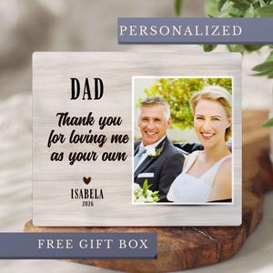 May include: A wooden photo frame with a picture of a smiling couple. The frame has the words "DAD, Thank you for loving me as your own" and the name "ISABELA" and the year "2026". The word "PERSONALIZED" is at the top.