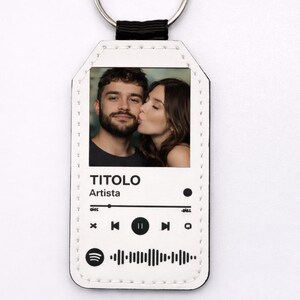 May include: White leatherette keychain with a silver ring. The keychain features a photo of a couple kissing, and a music player interface with the words "TITOLO" and "Artista". The design includes a Spotify code.