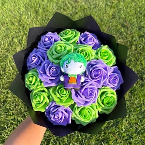 May include: A bouquet of artificial purple and lime green roses wrapped in black paper, featuring a small plush character with green hair, a white face, and a purple and orange suit positioned in the centre of the floral arrangement.