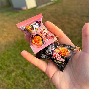 Pode incluir: Miniaturas de chaveiros de pacotes de ramen rosa e preto. O pacote rosa apresenta um personagem de desenho animado e o texto "Samyang". O pacote preto tem uma imagem de macarrão. Ambos têm argolas douradas para chaveiros.