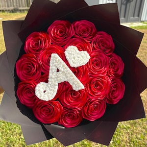Könnte beinhalten: Ein Bouquet aus roten Rosen, kreisförmig angeordnet und in schwarzes Papier gewickelt. Ein weißer Buchstabe "A" und ein weißes Herz sind auf den Rosen platziert. Das Bouquet befindet sich vor einem Hintergrund aus grünem Gras und einem Gebäude.