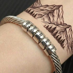 May include: A silver-toned cable bracelet featuring five cylindrical beads engraved with the words DAD, LOVE, BEST, HERO, and the date 2.2.2027. The bracelet is worn on a wrist near a mountain range tattoo.