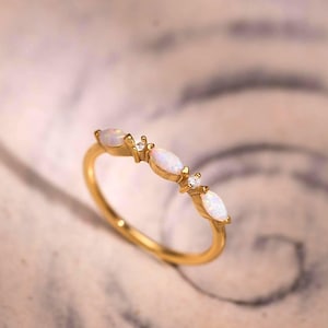 Puede incluir: Un anillo de oro con un diseño delicado. El anillo presenta tres piedras preciosas blancas de forma ovalada y dos piedras preciosas más pequeñas, redondas y transparentes. El anillo está sobre una superficie clara y arremolinada.