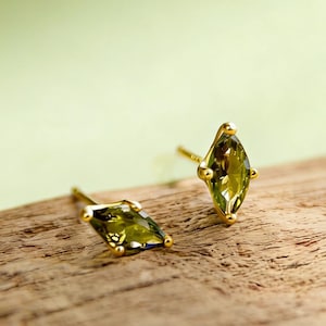 Puede incluir: Un par de pendientes de oro con gemas de talla marquesa verde oliva. Los pendientes están colocados sobre una superficie de madera con un fondo verde suave. Las gemas tienen una talla facetada.