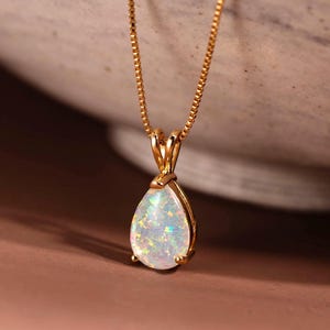 Op de afbeelding: Een goudkleurige ketting met een peervormige opaalhanger. De opaal heeft een witte basis met iriserende kleuren. De ketting is delicaat en de hanger is gezet in een gouden bezel.