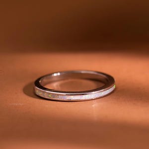 May include: A thin, polished silver-toned metal ring featuring a continuous, narrow inlay of iridescent white opal material running along the center of the band. The ring has a smooth, rounded profile and a reflective surface finish.