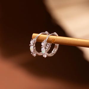 May include: A pair of small silver hoop earrings featuring a row of alternating clear round stones and oval-shaped iridescent white opals. The earrings are suspended from a thin, horizontal gold-toned metal rod, showcasing their circular design.
