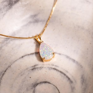 May include: A gold necklace with a pear-shaped opal pendant. The opal has a white and iridescent appearance, set in a gold bezel. The necklace is displayed on a marbled ceramic surface.