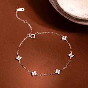 Könnte beinhalten: Ein filigranes Silberarmband mit fünf kleinen, funkelnden, blütenförmigen Anhängern. Das Armband hat eine feine Kette und einen sicheren Verschluss. Der Schmuck wird auf einer braunen Oberfläche präsentiert.