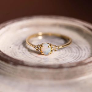 May include: A delicate gold ring featuring an oval opal center stone, flanked by small accent stones. The ring's band is thin and polished, and the opal has a milky white and iridescent appearance. The ring is set on a neutral-toned surface.