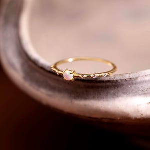 May include: A thin gold ring featuring a small, round, iridescent opal gemstone set in the center, flanked by two tiny, clear, sparkling accent stones on each side of the band, creating a delicate and refined piece of jewelry.