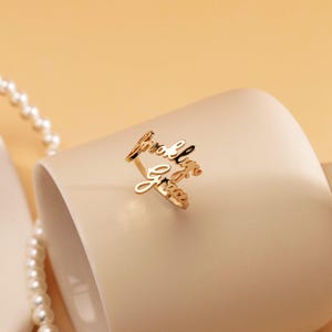 May include: A gold-toned metal ring featuring two stacked, cursive-style name plates that read Brooklyn and Grace. The ring is constructed with a thin, open-style band that connects the two script-lettered names in a layered, horizontal arrangement.