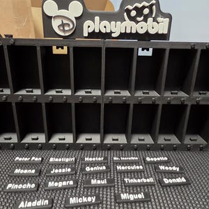 Pode incluir: Um expositor preto impresso em 3D com duas filas de compartimentos retangulares e uma placa superior com o logótipo da Disney e a palavra playmobil. Abaixo do expositor estão várias pequenas etiquetas retangulares pretas com texto branco.