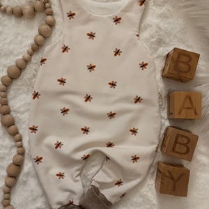 Könnte beinhalten: Ein cremefarbener Baby-Strampler mit einem sich wiederholenden Muster aus orangefarbenen und braunen Bienen. Der Strampler hat braune Bündchen und Schulterknöpfe. Holzblöcke schreiben "BABY". Eine Holzperlen-Girlande ist ebenfalls im Bild.