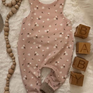 Könnte beinhalten: Ein Baby-Strampler in zartem Rosa, verziert mit einem zarten Blumenmuster in Weiß und Braun. Der Strampler hat ein weißes Innenfutter, braune Bündchen und Holzknopfverschlüsse. Holzblöcke schreiben "BABY".