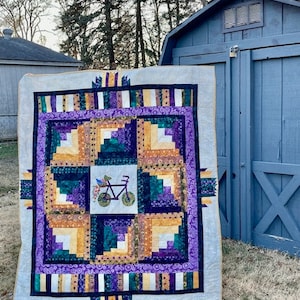 Puede incluir: Una colcha con un patrón de cabaña de troncos en tonos de morado, verde y dorado. El centro de la colcha presenta un diseño caprichoso de una bicicleta con flores.