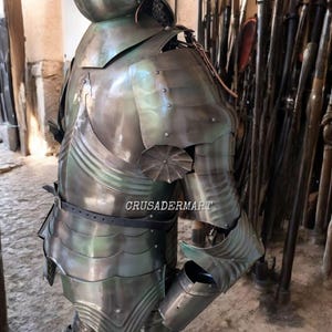 May include: A full suit of medieval armor, crafted from polished metal with a greenish hue. The armor includes a helmet, chest plate, shoulder guards, and arm and leg protection. The image shows the armor from the back, with the text "CRUSADERMART" visible.