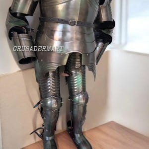 May include: A full suit of medieval armor in a dark, metallic finish. The armor includes a breastplate, arm and leg protection, and a belt. The text "CRUSADERMART" is visible on the chest. The armor is displayed on a wooden surface.