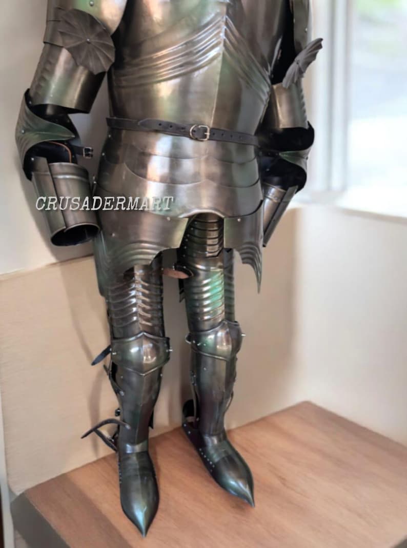 May include: A full suit of medieval armor in a dark, metallic finish. The armor includes a breastplate, arm and leg protection, and a belt. The text "CRUSADERMART" is visible on the chest. The armor is displayed on a wooden surface.