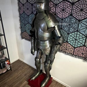 May include: Full suit of medieval armor, standing on a red base. The armor is made of metal and includes a helmet, chest plate, arm and leg protection, and gauntlets. The armor is displayed in a room with a patterned tapestry in the background.