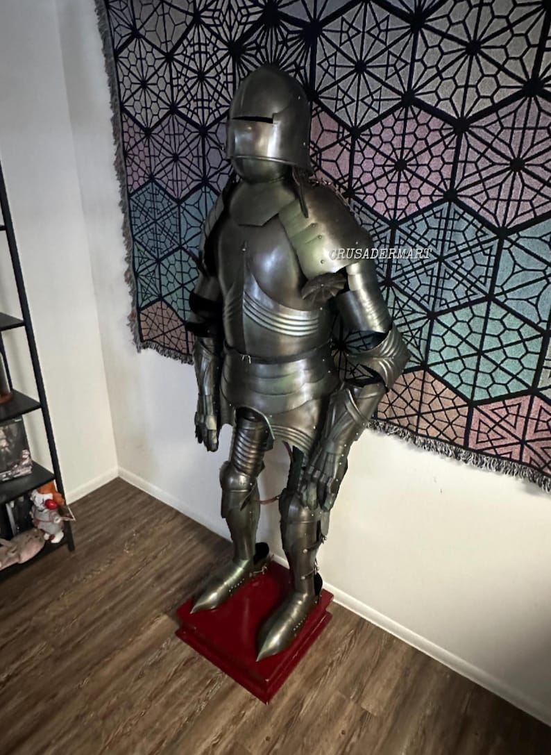 May include: Full suit of medieval armor, standing on a red base. The armor is made of metal and includes a helmet, chest plate, arm and leg protection, and gauntlets. The armor is displayed in a room with a patterned tapestry in the background.