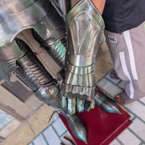 May include: Close-up of articulated metal armor gauntlets and leg armor. The gauntlets are silver-colored with detailed finger and wrist protection. The leg armor features pointed toe caps. The image is taken from a slightly elevated angle.