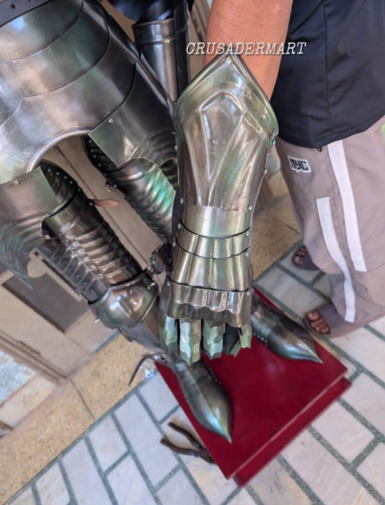 May include: Close-up of articulated metal armor gauntlets and leg armor. The gauntlets are silver-colored with detailed finger and wrist protection. The leg armor features pointed toe caps. The image is taken from a slightly elevated angle.