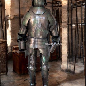May include: Full suit of medieval armor made of polished steel. The armor includes a helmet, chest plate, arm and leg protection, and is displayed in a stone room. The text "CRUSADERMART" is visible on the armor.