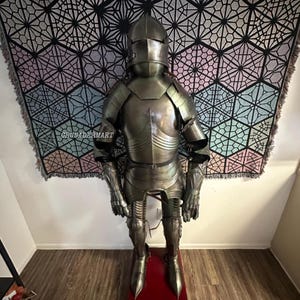 May include: Full suit of medieval armor, standing on a red base. The armor is metallic silver and includes a helmet, chest plate, and limb protection. A geometric patterned tapestry hangs in the background. The text "CRUSADERMART" is visible.