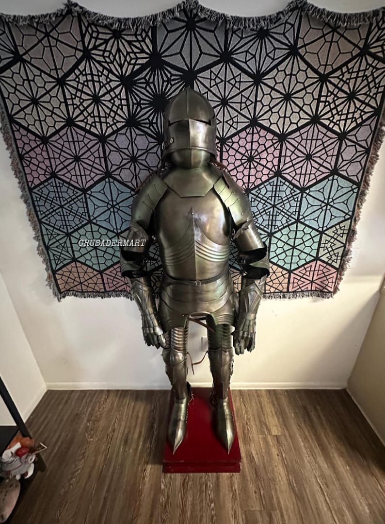 May include: Full suit of medieval armor, standing on a red base. The armor is metallic silver and includes a helmet, chest plate, and limb protection. A geometric patterned tapestry hangs in the background. The text "CRUSADERMART" is visible.