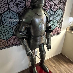 May include: Full suit of medieval armor, displayed on a red stand. The armor is a dark, metallic color with intricate detailing, including a helmet, chest plate, and leg guards. The background features a geometric patterned tapestry. The text "crusadermart" is visible.
