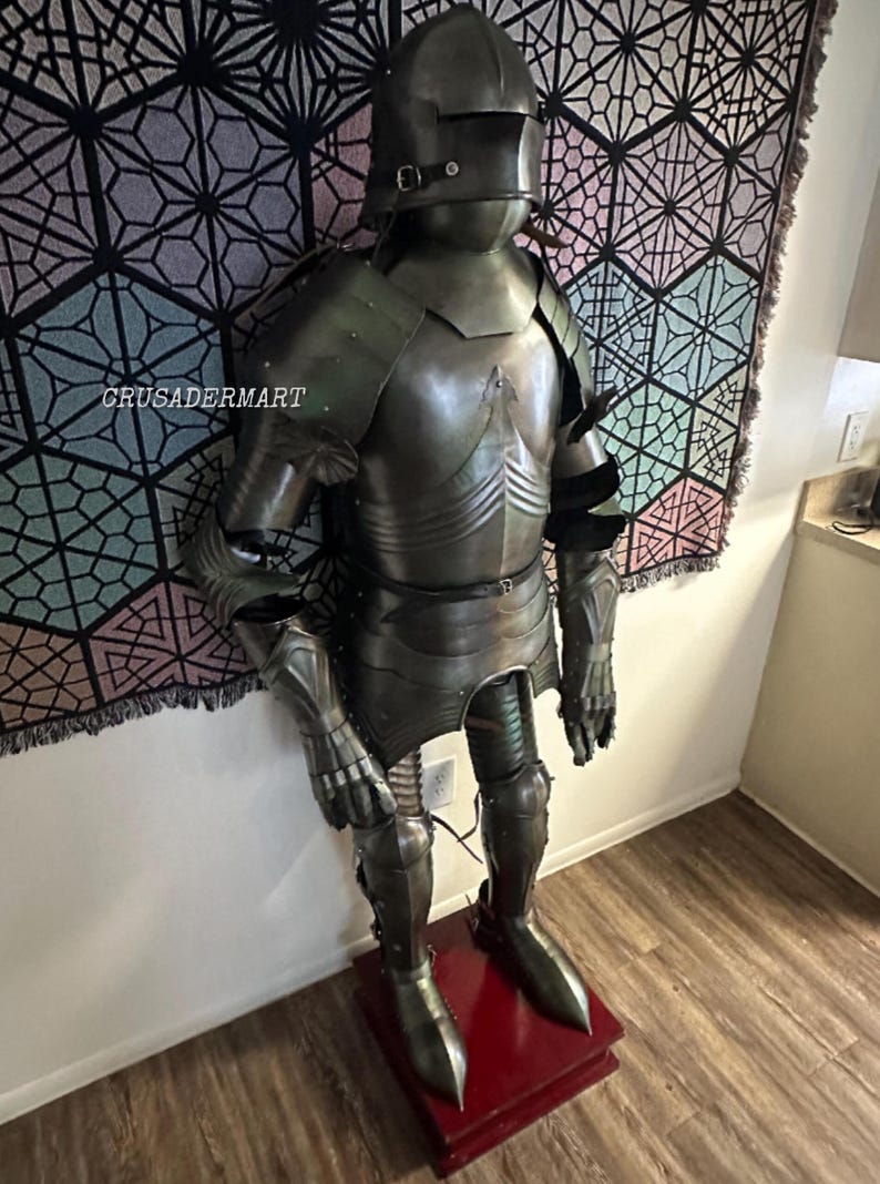 May include: Full suit of medieval armor, displayed on a red stand. The armor is a dark, metallic color with intricate detailing, including a helmet, chest plate, and leg guards. The background features a geometric patterned tapestry. The text "crusadermart" is visible.