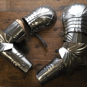 May include: A collection of polished steel arm armour, including gauntlets and elbow protection, displayed on a wooden surface. The armour features articulated plates and decorative elements, reflecting a medieval aesthetic.