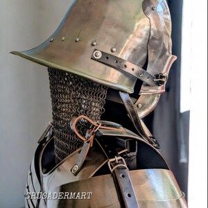 May include: A medieval-style metal helmet and chest armor set. The helmet has a wide brim and is riveted. The chest armor is secured with leather straps. A chainmail collar is visible. The text "CRUSADERMART" is printed on the chest armor.