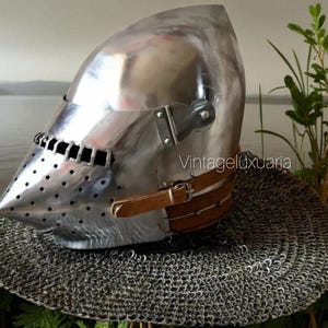 Puede incluir: Un casco de metal pulido con visera puntiaguda, detalles de remaches y una correa de cuero con hebilla. El casco descansa sobre una cofia de cota de malla. Fondo con agua y vegetación.