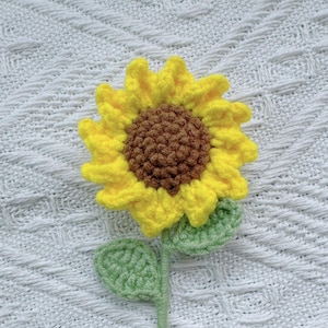 May include: A handmade crochet sunflower with yellow petals, a brown center, green leaves, and a green stem. The sunflower is placed on a white, textured background. This floral craft item is ideal for decorative purposes.