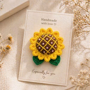 Könnte beinhalten: Eine handgefertigte gehäkelte Sonnenblume auf einer Karte mit dem Text "Handmade with love" und "Especially for you". Die Sonnenblume hat gelbe Blütenblätter, eine braune und rosa Mitte und grüne Blätter. Die Karte liegt auf einer beigen Oberfläche.