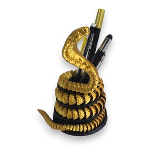 May include: A gold-coloured snake-shaped desk organiser. The coiled body forms the base, with the snake's head rising to the top. Black pens and a stylus are visible inside. The organiser is designed to hold pens and pencils.
