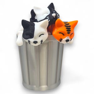 May include: A silver bin filled with three cat ornaments. One is black and white, another is black and white with spots, and the third is orange with black stripes. All cats have their eyes closed.
