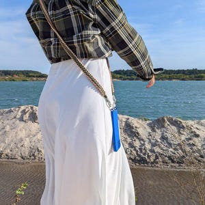 Puede incluir: Una persona con una camisa a cuadros verde y negro y pantalones blancos. Una funda azul para teléfono cuelga de una correa trenzada. El fondo presenta un lago y un cielo azul.