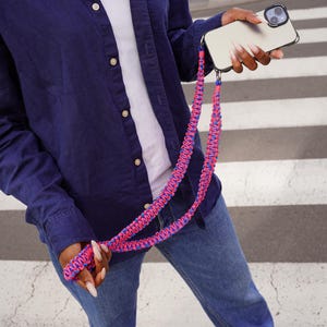 Puede incluir: Un smartphone plateado con una funda negra y un cordón trenzado rosa y azul. El cordón lo sostiene una persona que lleva una camisa abotonada azul marino, una camiseta blanca y vaqueros azules.