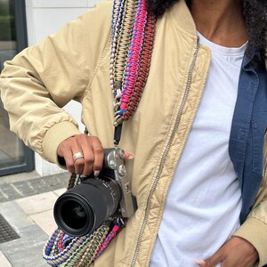 May include: A person wearing a beige jacket with a multi-colored braided camera strap. A black camera with a silver lens is attached to the strap.