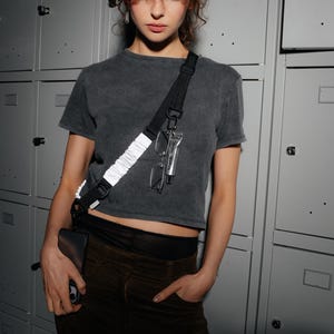 May include: A dark gray t-shirt with a black strap across the body, holding glasses and a tube of cream. The person is wearing brown pants and holding a black wallet. The background is a series of gray lockers.
