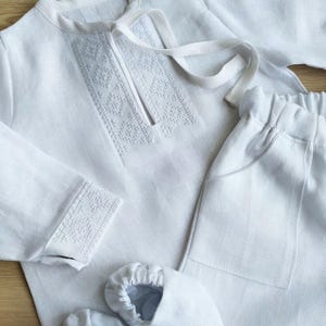 Puede incluir: Un conjunto de bebé de lino blanco con detalles bordados. El conjunto incluye una camisa de manga larga, pantalones y botines.