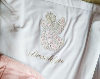 May include: White blanket with an embroidered angel design in pink and beige. The name "Brooklyn" is embroidered below the angel in a matching beige thread. The blanket is folded, with a glimpse of a pink fabric visible.