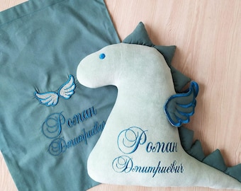 May include: A green fabric bag with a blue embroidered design of wings and the name "Roman Demitriyev".  A light green plush dinosaur toy with blue embroidered wings and the name "Roman Demitriyev" is lying next to the bag.